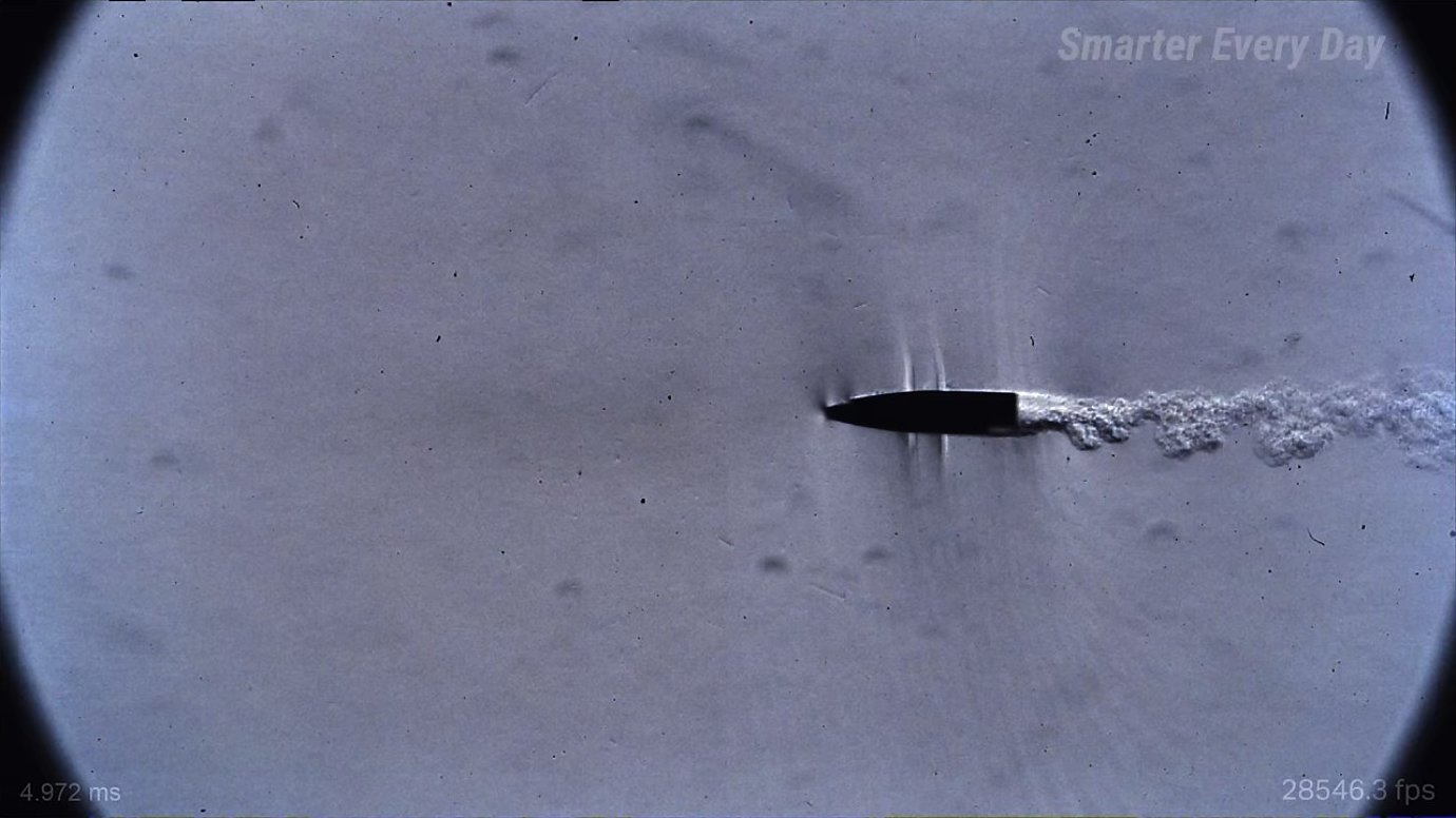 A bullet captured at the instant it pierces through its own muzzle blast. The overlapping shock waves from projectile and propellant gases create a richly structured density field.