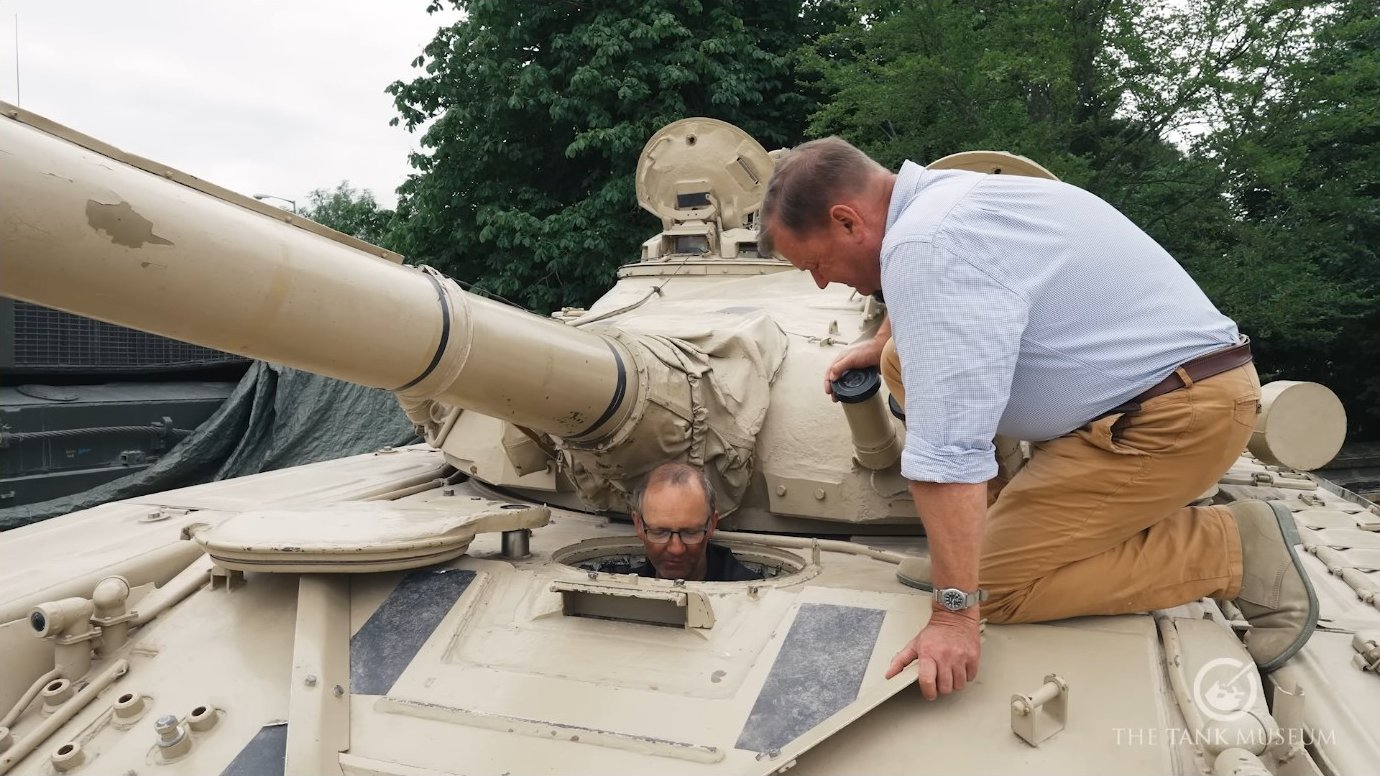The commander and gunner inspect the turret top. The commander's forward-opening hatch provides weather protection and a degree of cover, though it limits overhead clearance when open.