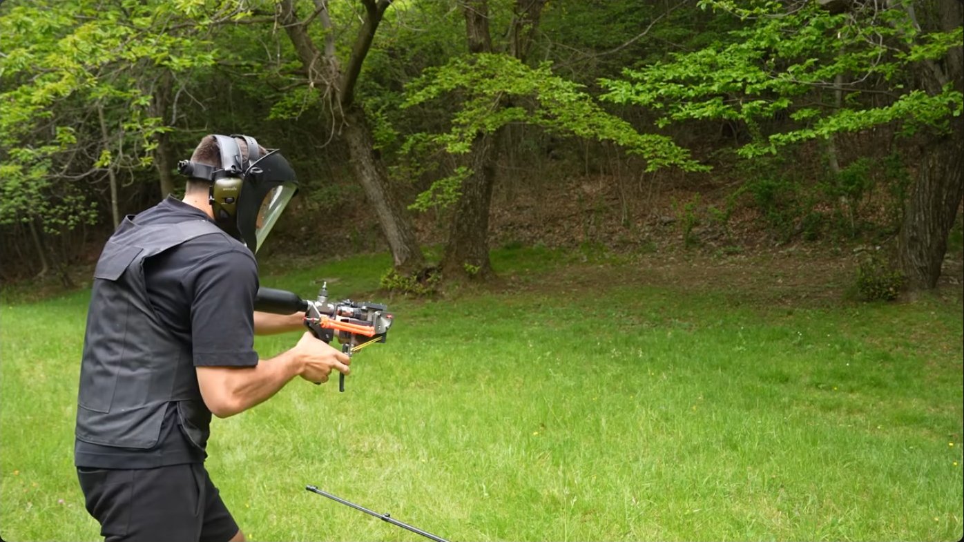 Test firing the pneumatic cannon in an open field, demonstrating the proper stance and protective equipment required when operating high-pressure air systems. Note the substantial barrel elevation from recoil.